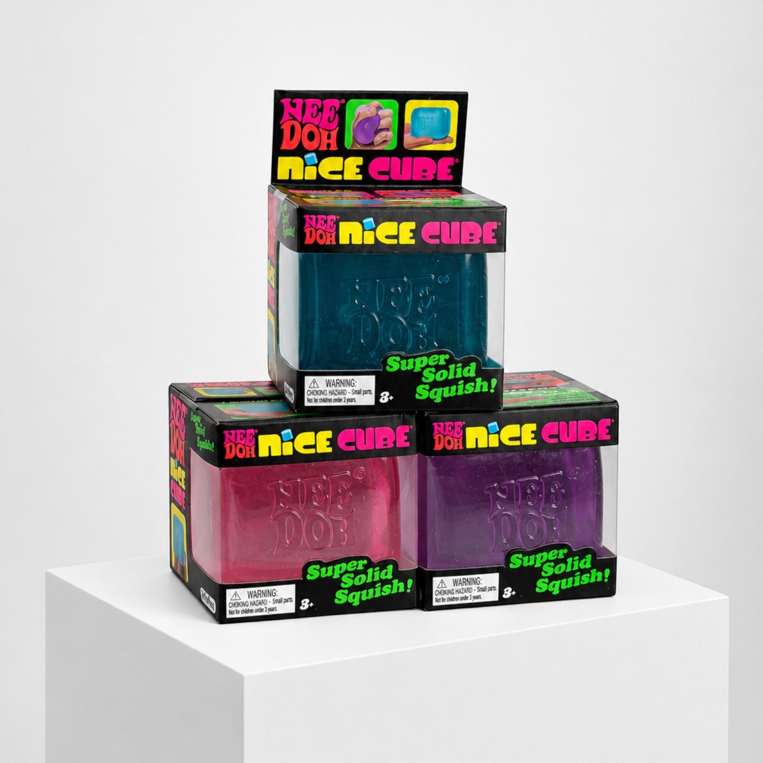 Squish Planet NeeDoh Nice Cube fidget toys in blue, pink, and purple boxes stacked on a white pedestal.