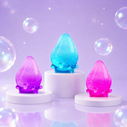 Squish Planet NeeDoh Dream Drop fidget toys in purple, blue, and pink on white pedestals with bubbles.