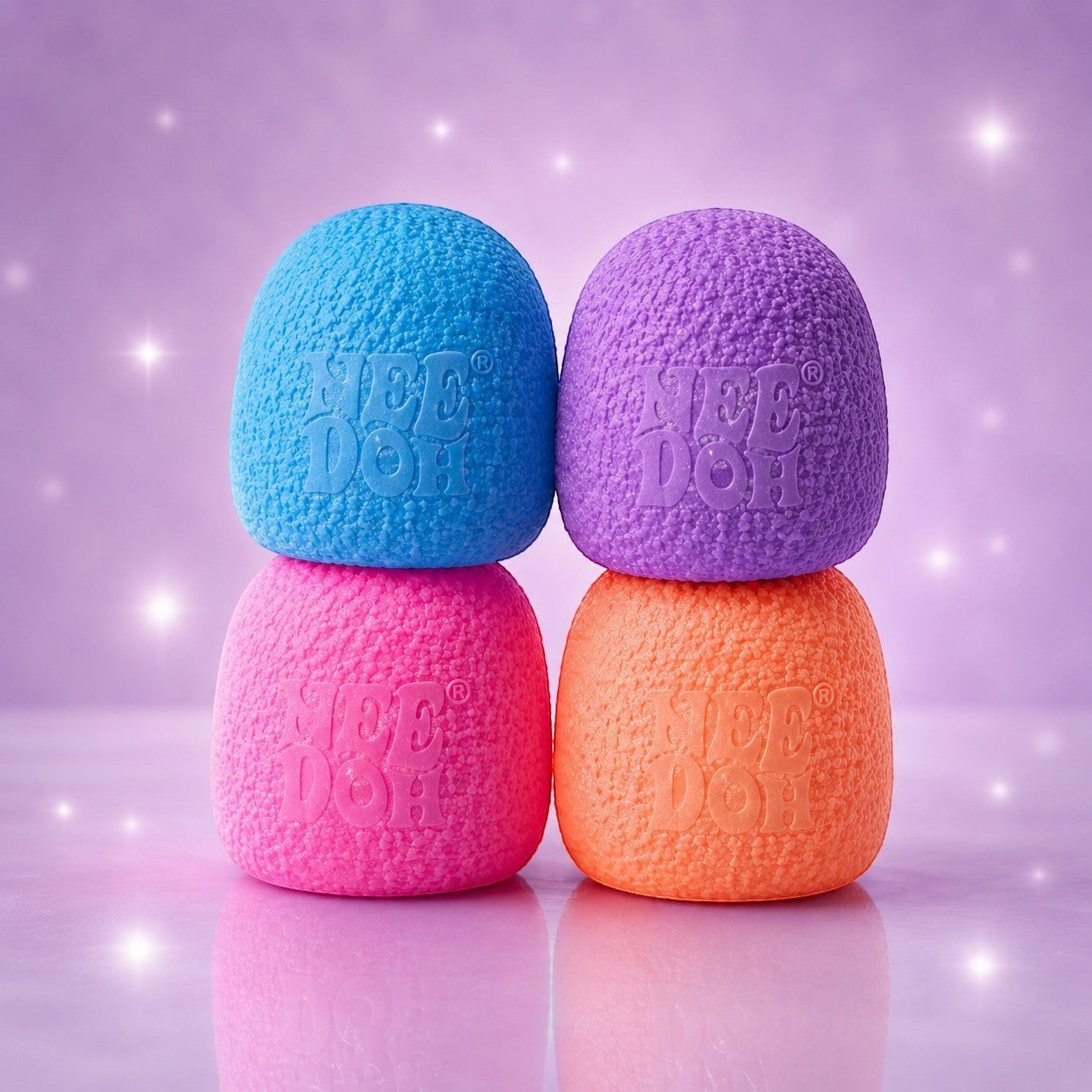 Squish Planet NeeDoh Gumdrop fidget toys in blue, purple, pink, and orange stacked on a sparkly purple background.